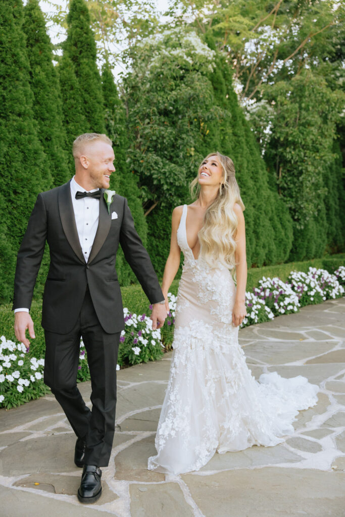 A newlywed couple holding hands and walking on a path at a Russos on the Bay wedding.