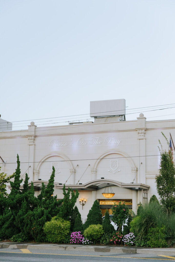 The exterior sign for Russo's On The Bay, a wedding venue, is visible above green bushes and flowers.