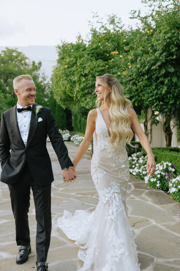 A newlywed couple holds hands and walks on a path at their Russos on the Bay wedding.