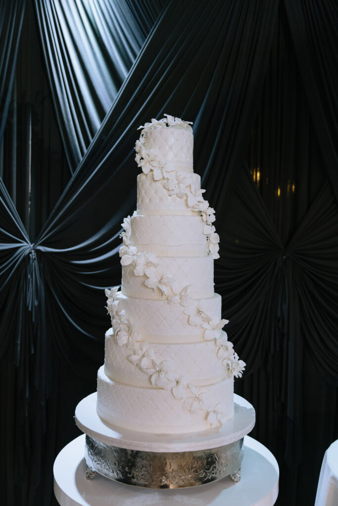 A towering white wedding cake with floral accents sits on a silver stand at Russos on the Bay.