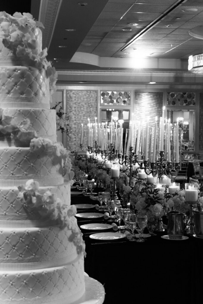 A multi-tiered wedding cake with white flowers next to a candlelit reception table at Russos on the Bay.