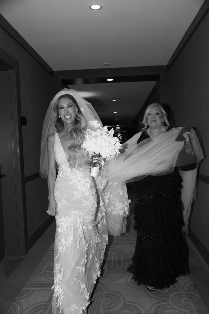 A bride in a white gown and a woman in a black dress walk down a hotel hallway at a Russos on the Bay wedding.