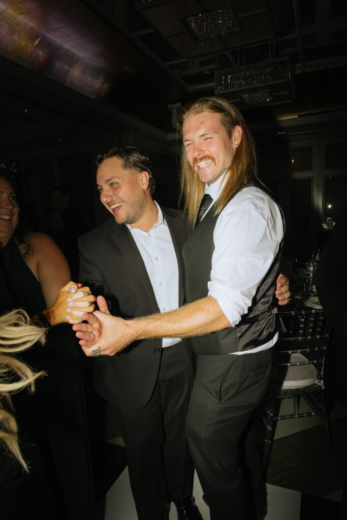 Two men laugh and dance together at a glamorous wedding reception at Russos on the Bay.