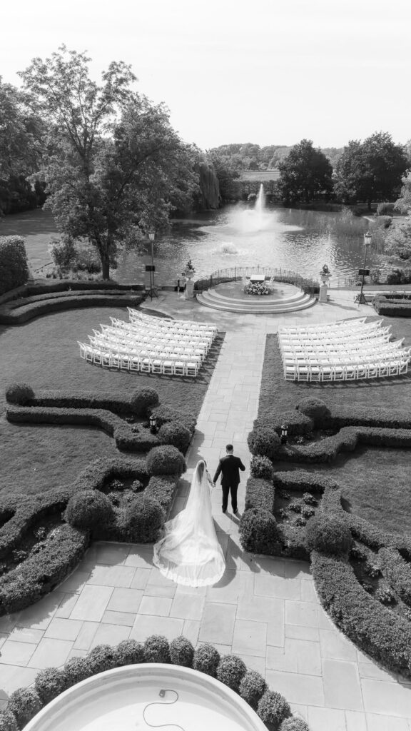 A newlywed couple walks hand-in-hand towards a formal garden wedding ceremony at Park Chateau.