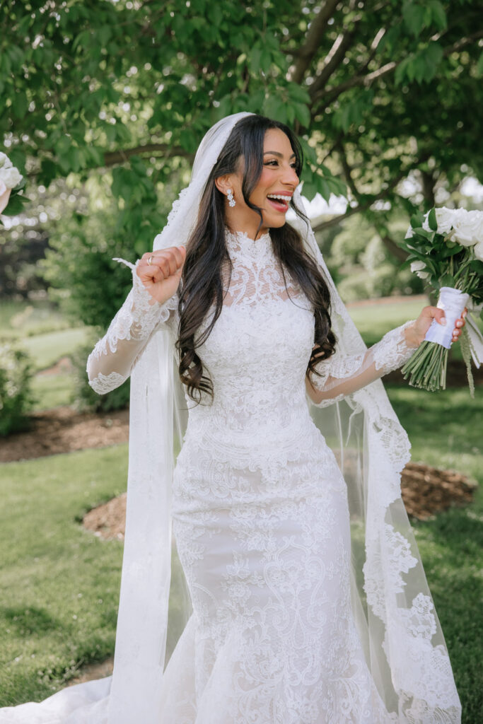 A bride in a white lace wedding dress laughs while holding a white rose bouquet at Park Chateau.