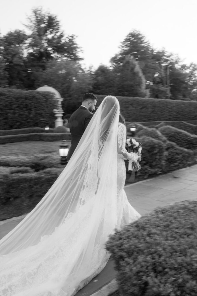 A groom and a bride walking, with the bride's long veil flowing behind her at Park Chateau, New Brunswick, New Jersey.