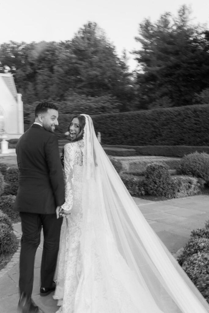 A groom and a bride in a lace dress and long veil walk, smiling over shoulders at Park Chateau.