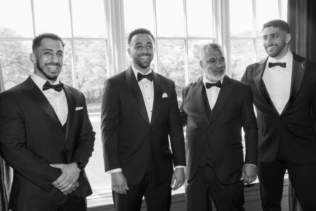 Men posing in tuxedos by a large window at a Park Chateau New Brunswick, New Jersey wedding.