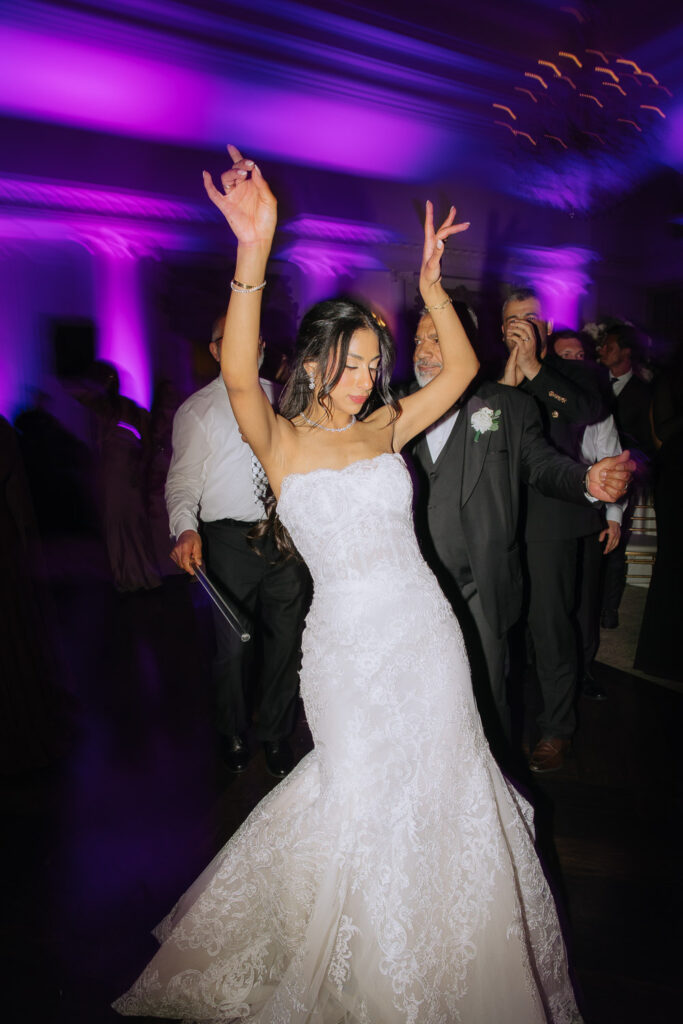 A bride dances with arms raised in her white lace wedding dress at Park Chateau in New Brunswick, NJ.