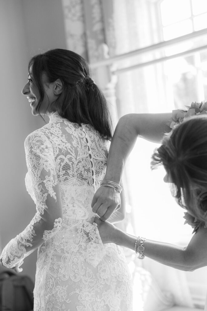 A woman smiles as another woman helps button up the back of her lace wedding dress at Park Chateau.