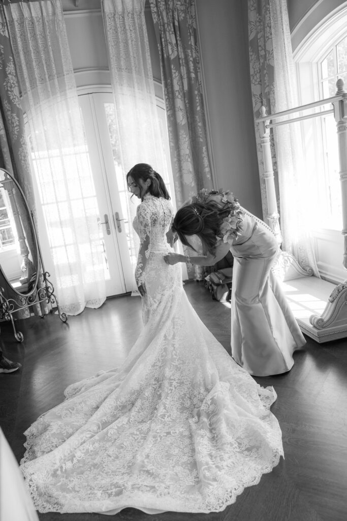 A woman fastens the back of a bride's long-sleeved lace wedding dress at Park Chateau New Brunswick.