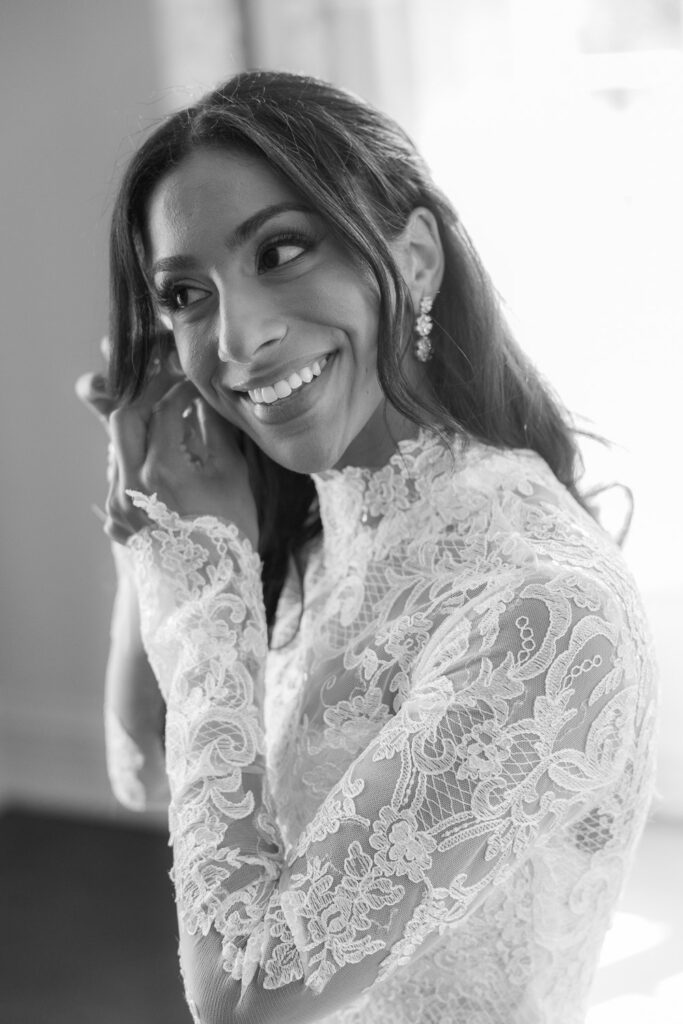 A smiling bride wearing a lace wedding dress puts on an earring at Park Chateau, New Brunswick, NJ.