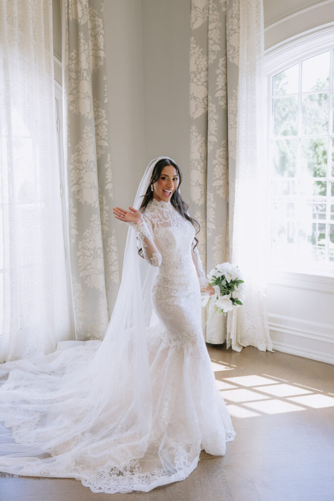A bride in a white lace wedding dress smiles, holding a white bouquet at Park Chateau New Brunswick.
