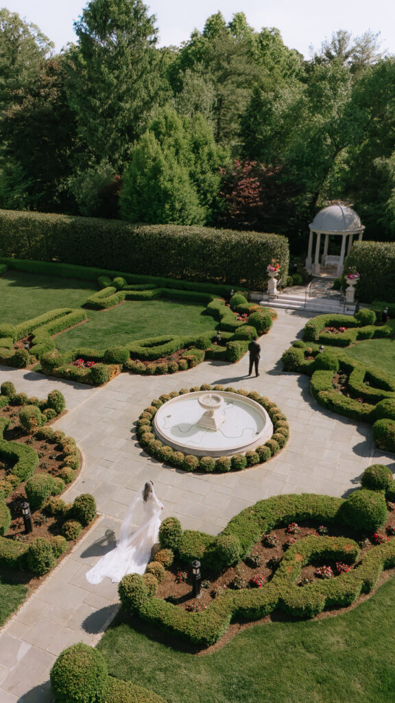 A bride walks towards her groom on a paved path in the formal gardens at Park Château Estate & Gardens.