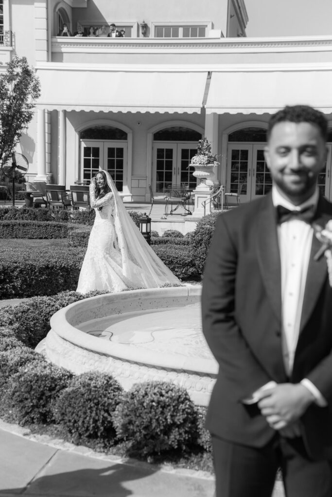A groom awaits his bride, who is seen approaching with a long train at Park Château Estate & Gardens.